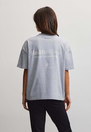 Kvinde med brunt hår står med ryggen til iført en grå T-shirt med teksten "Health Wellness Feel good. Live well. Be you." på ryggen.
