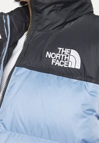 Czarna i jasnoniebieska ocieplana kamizelka z zamkiem błyskawicznym, wysokim kołnierzem i haftowanym białym logo "The North Face". Gładka faktura.