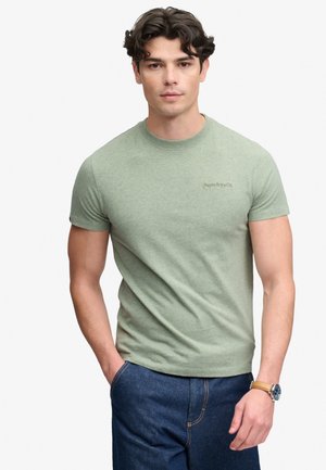 Giovane uomo con capelli scuri che indossa una maglietta Superdry verde chiaro e jeans blu, con una mano in tasca e un orologio da polso al polso sinistro.
