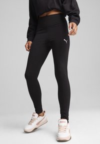 Czarne legginsy wykonane z elastycznej tkaniny, z wysokim stanem i białym logo Puma po lewej stronie, w połączeniu z białymi sneakersami.