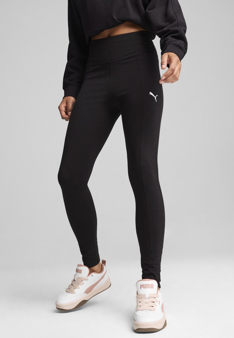 Czarne legginsy wykonane z elastycznej tkaniny, z wysokim stanem i białym logo Puma po lewej stronie, w połączeniu z białymi sneakersami.