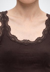 Gros plan d'une personne portant un débardeur marron côtelé avec une bordure en dentelle le long de l'encolure et des épaules.