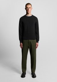 Zwarte sweatshirt met geribbelde halslijn en lange mouwen, gecombineerd met olijfgroene broek. Model draagt zwarte veterschoenen met gestructureerde zolen.