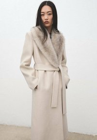 Abrigo de lana beige con un amplio cuello de piel, cintura ajustada, mangas largas y dos bolsillos frontales, con un cinturón de lazo para cierre.