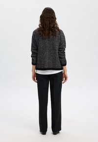 Črno-bel pleteni pulover z vzorcem ribje kosti in zavihanim rokavom, oblečen čez belo majico, kombiniran s črnimi širokimi hlačami.