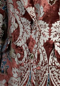 Chaqueta de brocado en burdeos profundo con patrón floral plateado. Presenta botones metálicos y una tela texturizada, aumentando la profundidad visual.