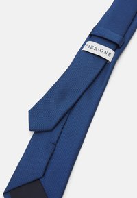 Corbata texturizada azul con punta afilada, que presenta una etiqueta visible que dice "PIER·ONE". La tela tiene un patrón tejido.