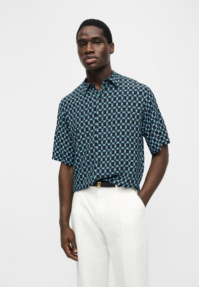 Άνδρας με μαύρο πουκάμισο κοντού μανικιού με ανοιχτό μπλε γεωμετρικό σχέδιο και λευκό παντελόνι, στέκεται με το ένα χέρι στην τσέπη.