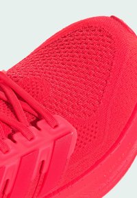 Helles rotes Sportschuh mit einem strukturierten Mesh-Obermaterial, verstärktem Fersenbereich und breiten Schnürsenkeln, einer glatten Gummisohle und Markenakzenten.