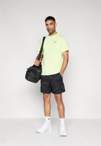 Λαχανί πράσινη μπλούζα Nike, μαύρα σορτς καμουφλάζ, λευκά αθλητικά παπούτσια και μαύρη τσάντα με ιμάντα ώμου. Ομαλή υφή, casual αθλητικός σχεδιασμός.