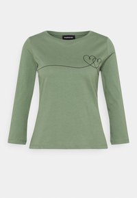 Vihreä pitkähihaisessa paidassa on pyöreä pääntie, ja etuosassa vasemmassa ylärintakehässä on mustasta, jatkuvasta viivasta muodostettu kaksi toisiinsa kytkettyä sydäntä.