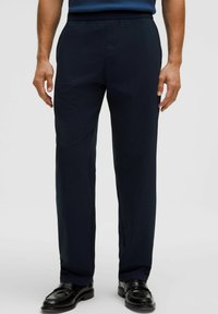 Hombre vistiendo pantalones de vestir negros y zapatos de cuero negros, de pie contra un fondo claro y sencillo.