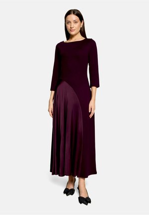 Femme portant une longue robe violet foncé à manches trois-quarts et des chaussures noires pointues avec des nœuds, debout devant un fond uni.