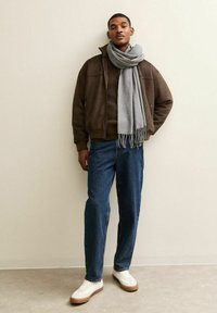 Bruine bomberjack over een donkere trui, blauwe jeans en witte sneakers. Een grijze sjaal hangt om de nek en creëert een gelaagde look.