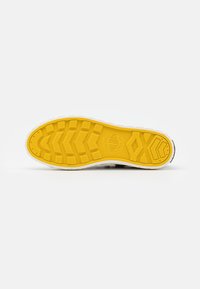 Semelle de chaussure avec une bande de roulement jaune, présentant des motifs géométriques et un logo circulaire au centre, sur un fond blanc uni.