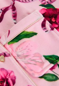 Rosa Fleece mit Blumenmuster und einer Tasche, die mit einem herzförmigen goldenen Akzent versehen ist; glatte Textur und lebendige Farben von Blumen und Blättern.