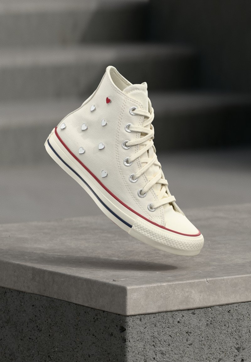 Baskets montantes blanches avec des clous en forme de cœur rouges et blancs, des lacets blancs et des rayures rouges et bleues sur la semelle, flottant au-dessus d'un bloc de béton gris.