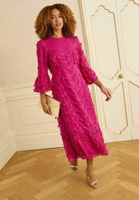 Fuksia keskmise pikkusega kleit tekstuurse kangaga, volangikeste ja kihtide disainiga. Täiendatud metallik-kuldsest käekotist ja rihmikestest kontsakingadest.