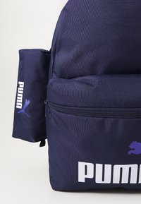 Mørkeblå ryggsekk med teksturert stoff, med en frontlomme, glidelås-lukking og hvit Puma-logo. Sidevott for ekstra oppbevaring.