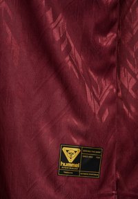 Viinapunane spordikangas peente geomeetriliste mustritega ja must-kuldne hummel spordirõivaste silt, millel kirjas „Serving the game since 1923.“