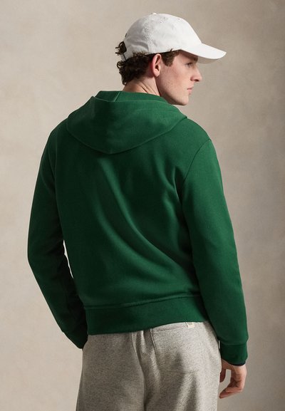 Mees, kellel on valge kork, roheline kapuutsiga dressipluus ja hallid dressipüksid, vaadates küljepealt lihtsa beežika tausta vastu.