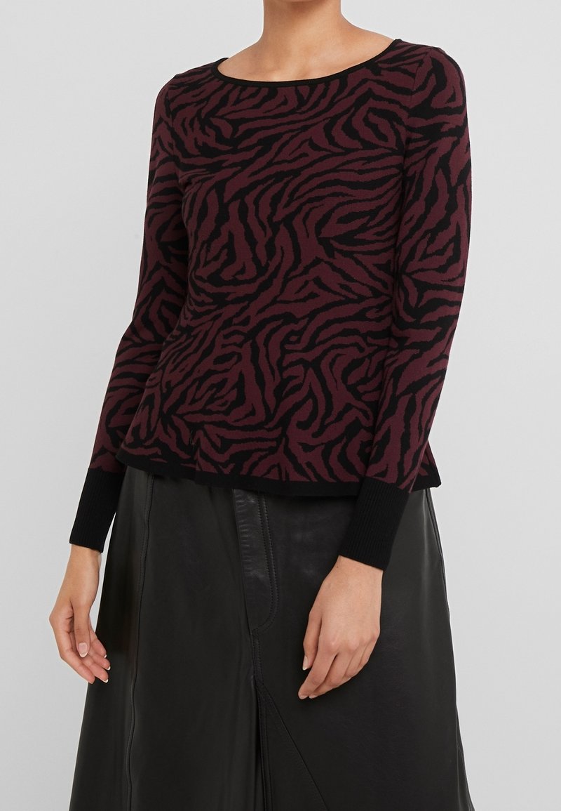 Femme portant un haut à manches longues imprimé zèbre bordeaux et noir avec des poignets noirs et une jupe en cuir noire.