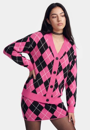 Jonge vrouw die een roze en zwarte argylevest en bijpassende rok draagt, poserend met één hand op haar heup tegen een effen achtergrond.