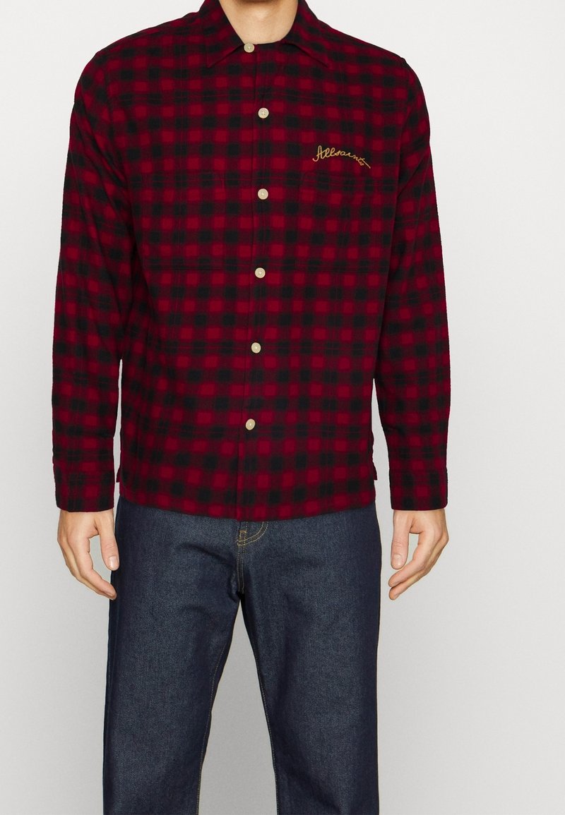 Homme portant une chemise à carreaux rouge et noire avec un logo brodé jaune sur la poitrine gauche, associée à un jean bleu foncé.