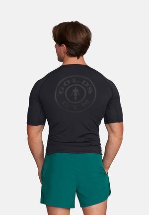 Mann mit kurzem braunem Haar, der ein eng anliegendes schwarzes Gold's Gym T-Shirt und türkisfarbene Sportshorts trägt, von hinten gesehen mit den Händen in den Taschen.