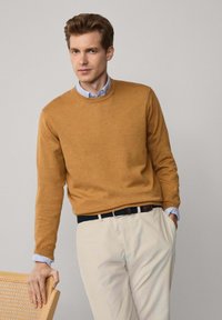 Sweter w kolorze musztardowym z okrągłym dekoltem, długimi rękawami i ściągaczami, noszony na jasnoniebieskiej koszuli z kołnierzykiem i beżowych spodniach.