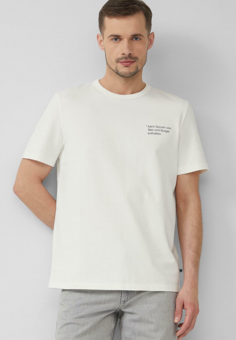 Witte katoenen t-shirt met korte mouwen, ronde hals en zwarte tekst op de linkerkant van de borst, met een humoristisch bericht over ingrediënten.