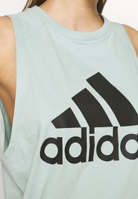Top verde chiaro senza maniche in cotone, con un grande logo Adidas nero sul petto, design senza maniche e vestibilità comoda.