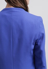 Blazer a medida de un azul real con una textura suave, solapas muestradas y costuras contorneadas. La vista trasera destaca un ajuste estructurado y líneas limpias.