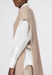 Beige geripptes Strickmuster ohne Ärmel über einem weißen Hemd mit Knopfleiste. Features Seitenschlitze und einen Rundhalsausschnitt. Strukturiertes, modernes Aussehen.