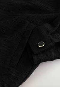 Material negru texturat cu un buzunar dreptunghiular și un clapetă fixată de un nasture negru rotund cu margine aurie.