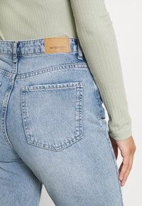 Jeans de mezclilla azul claro con cintura alta, que presentan una parche de cuero marrón claro etiquetado como "jeans perfectos". La tela de algodón tiene una textura sutil.