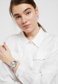 Silver wristwatch with a round face, minimalistic design, mesh band, and three simple hour markers. Worn on a wrist against a white shirt.