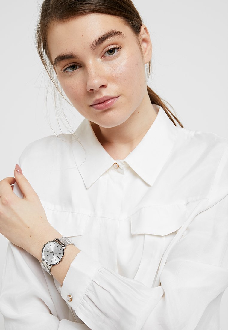 Silver wristwatch with a round face, minimalistic design, mesh band, and three simple hour markers. Worn on a wrist against a white shirt.