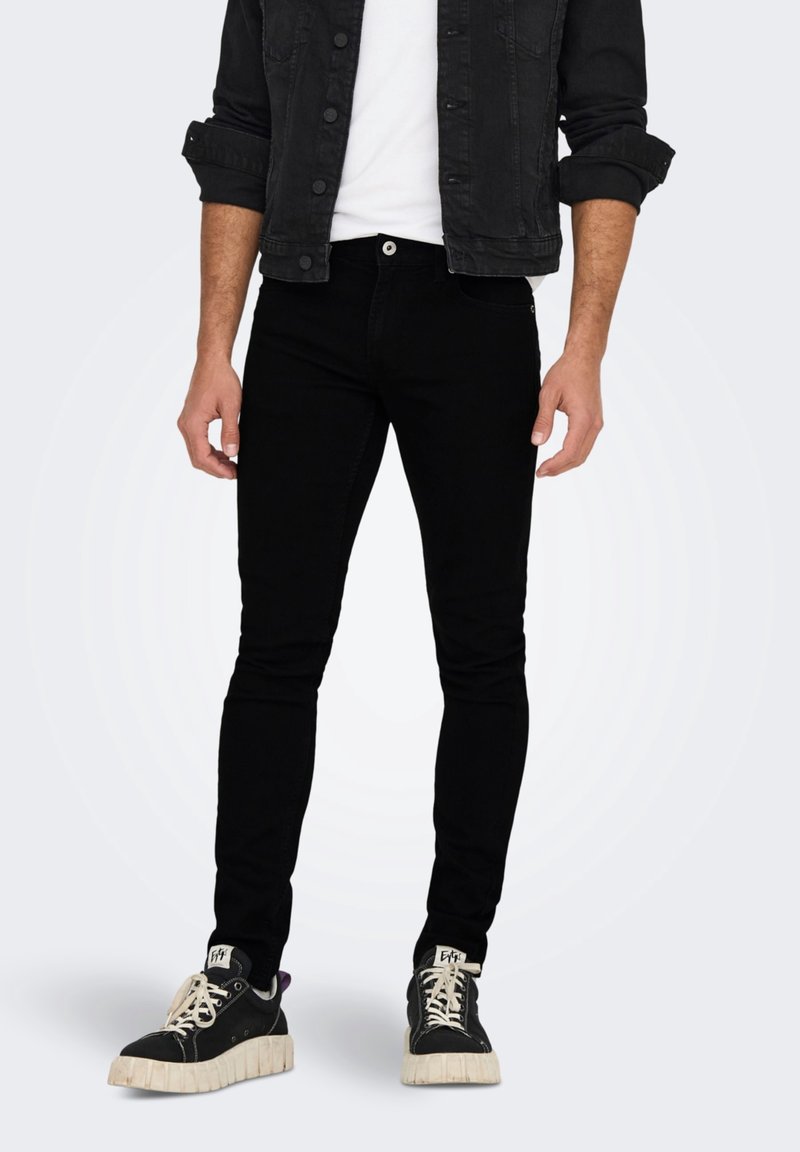 Jeans negros de corte slim hechos de mezclilla, combinados con una camiseta blanca y una chaqueta de mezclilla negra. El calzado presenta zapatillas negras con suelas gruesas.