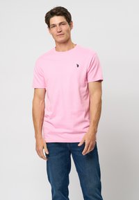 Man staat tegen een effen achtergrond, draagt een roze T-shirt met korte mouwen met een klein zwart logo en een blauwe spijkerbroek.