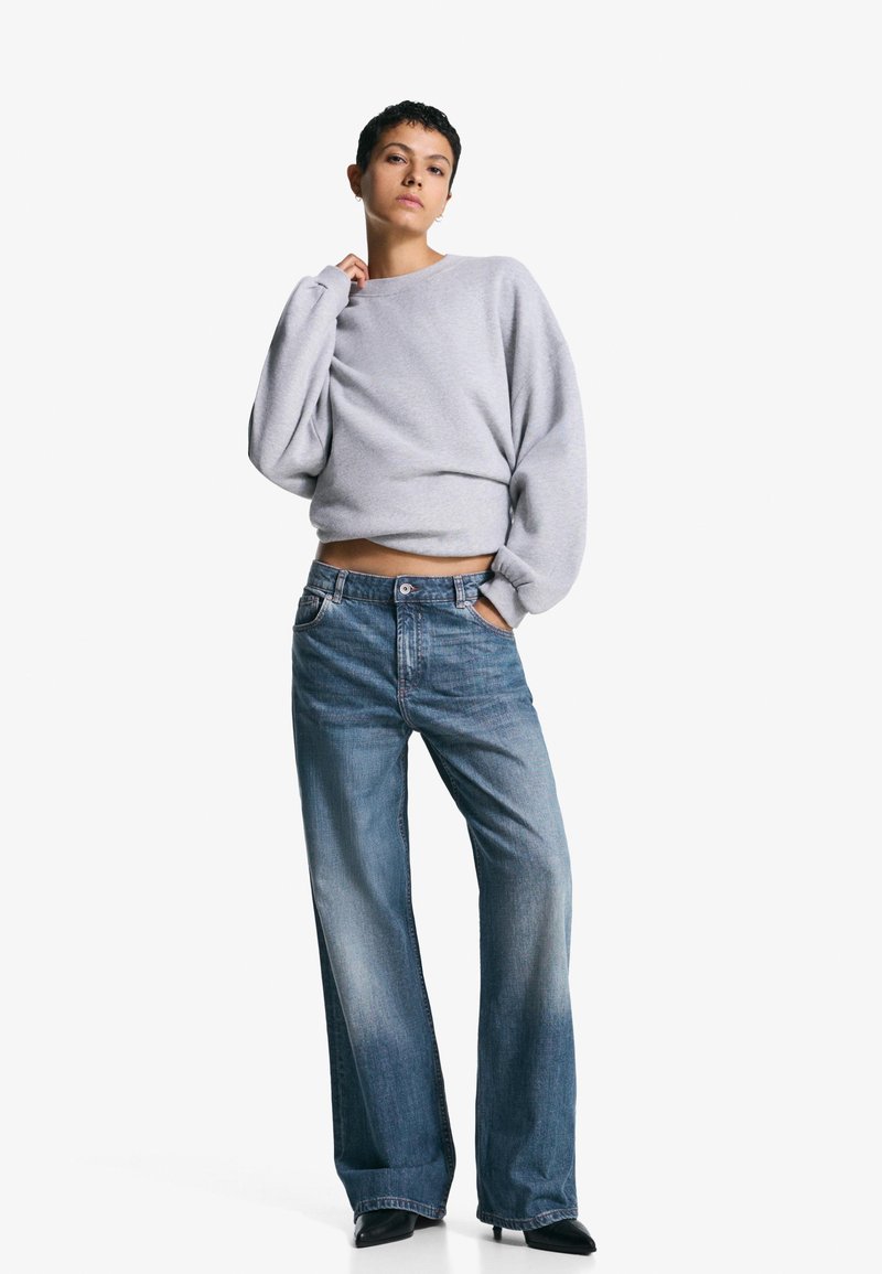 Siv sweatshirt z ohlapnim krojem in dolgimi rokavi, povezan z svetlo modrimi širokimi hlačami, ki imajo standardno zasnovo petih žepov in kovinski gumb.