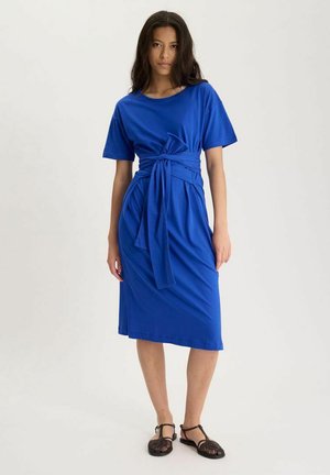 Femme aux longs cheveux foncés portant une robe midi bleu vif à manches courtes avec une ceinture nouée à la taille, associée à des sandales noires à bouts ouverts.