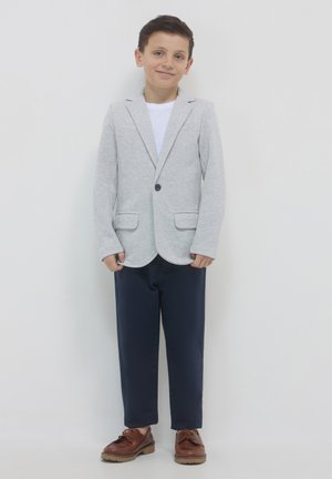 Hellgrauer Blazer mit Schneppkragen, zwei aufgesetzten Taschen und einem Knopf. Kombiniert mit navyfarbigen Hosen und braunen Schnürschuhen.