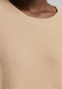 Gros plan sur une personne portant un pull en maille beige texturée avec un col rond côtelé.