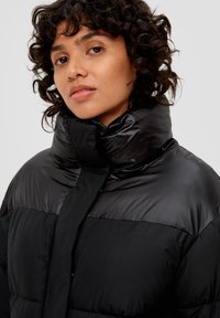 Schwarze Daunenjacke mit hohem Kragen, glänzender Oberfläche und gestepptem Muster. Das Design umfasst einen Vorderverschluss.
