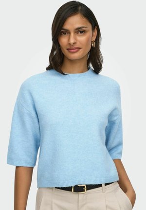 Žena s tmavými vlasmi dĺžky po plecia, ktorá má zlaté kruhové náušnice, svetlomodrý pulóver s krátkym rukávom a béžové nohavice s čiernym opaskom.