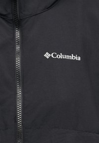 Ύφασμα μαύρου μπουφάν με κεντρικό φερμουάρ και λευκό λογότυπο Columbia τυπωμένο στην δεξιά πλευρά του στήθους.