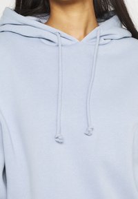 Sweat à capuche bleu clair en tissu doux, présentant un col en V, des cordons de serrage et une texture lisse. Aucun motif ni ornement visible.