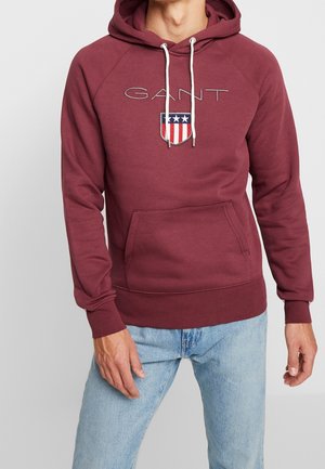 Sudadera color marrón burdeos con un bolsillo frontal tipo pouch, cordón ajustable, puños acanalados y un logo bordado con acentos en rojo, blanco y azul.