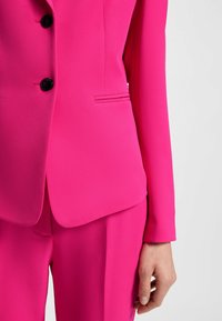 Costume ajusté fuchsia comprenant une veste à un bouton avec des boutons noirs et une poche élégante ; tissu lisse et texturé ; pantalon ajusté.